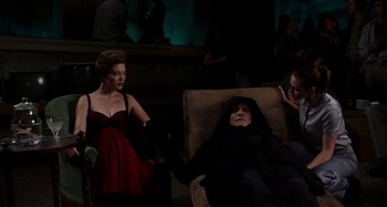 Movie still from “Crimes of the Future” (2022), directed by David Cronenberg – A woman in a red dress sitting next to a man in a black robe; Wide shot, High angle