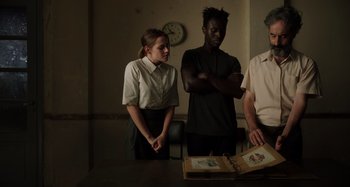 Movie still from “Crimes of the Future” (2022), directed by David Cronenberg – A group of people standing around a table; Medium shot, Over the shoulder angle