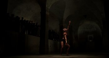 Movie still from “Crimes of the Future” (2022), directed by David Cronenberg – A man in black shorts and a black shirt is in a dark room; Extreme Wide shot, Low angle