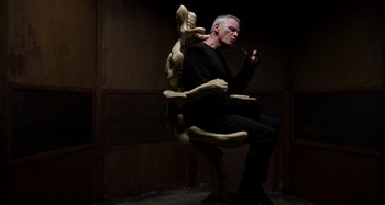 Movie still from “Crimes of the Future” (2022), directed by David Cronenberg – A man sitting in a chair with a dog statue on his lap; Medium shot, Low angle