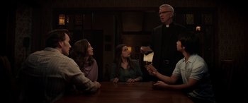 Movie still from “The Conjuring: The Devil Made Me Do It” (2021), directed by Michael Chaves – A group of people sitting at a table with a priest; Medium shot, Over the shoulder angle