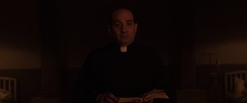 Movie still from “The Conjuring: The Devil Made Me Do It” (2021), directed by Michael Chaves – A man wearing a priest's outfit holding a book; Medium shot, Low angle