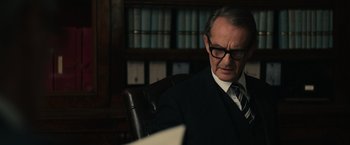 Movie still from “The Courier” (2020), directed by Dominic Cooke – An older man sitting in a chair in front of a book case; Medium shot, Over the shoulder angle