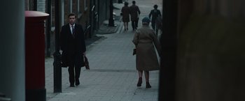 Movie still from “The Courier” (2020), directed by Dominic Cooke – An older woman walking down a sidewalk with a man in the background; Wide shot, Over the shoulder angle