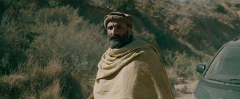 Movie still from “The Covenant” (2023), directed by Guy Ritchie – A man with a beard wearing a blanket and a hat; Medium shot, Over the shoulder angle