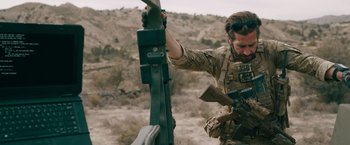 Movie still from “The Covenant” (2023), directed by Guy Ritchie – A man holding a rifle in his hand while standing next to a machine gun; Medium shot, Over the shoulder angle