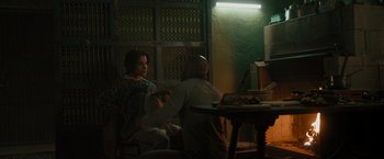 Movie still from “The Covenant” (2023), directed by Guy Ritchie – Two people sitting in a room with a table and a lamp; Medium shot, Over the shoulder angle