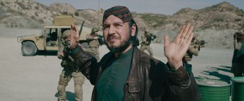 Movie still from “The Covenant” (2023), directed by Guy Ritchie – A man in a leather jacket and a hat is holding his hands in the air; Medium shot, Over the shoulder angle