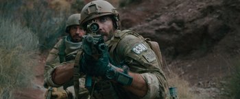 Movie still from “The Covenant” (2023), directed by Guy Ritchie – A man with a rifle aiming at something in his hand; Medium shot, Over the shoulder angle