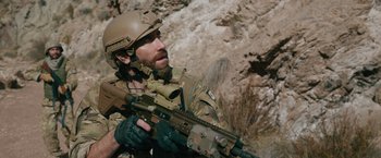 Movie still from “The Covenant” (2023), directed by Guy Ritchie – A man with a helmet on holding a rifle; Medium shot, Over the shoulder angle