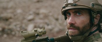 Movie still from “The Covenant” (2023), directed by Guy Ritchie – A man with a beard and a helmet is holding a rifle; Close Up shot, Over the shoulder angle