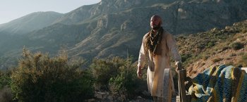 Movie still from “The Covenant” (2023), directed by Guy Ritchie – A bald man in a white robe and a scarf is standing on a hill; Wide shot, Low angle