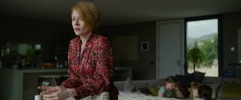 Movie still from “The Covenant” (2023), directed by Guy Ritchie – A woman in a red and black floral shirt sitting in a living room; Medium shot, Low angle