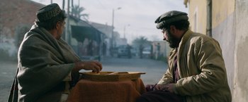 Movie still from “The Covenant” (2023), directed by Guy Ritchie – Two men sitting at a table in front of a street; Medium shot, Over the shoulder angle
