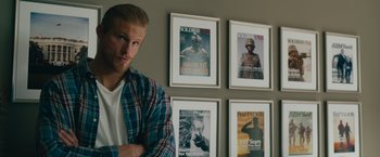Movie still from “The Covenant” (2023), directed by Guy Ritchie – A man standing in front of a wall with four framed magazines; Medium shot, Over the shoulder angle