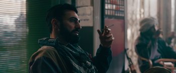 Movie still from “The Covenant” (2023), directed by Guy Ritchie – A bearded man holding a cigarette in his right hand; Close Up shot, Over the shoulder angle