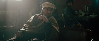 Movie still from “The Covenant” (2023), directed by Guy Ritchie – A man with a beard and a hat is smoking a cigarette; Medium shot, Over the shoulder angle