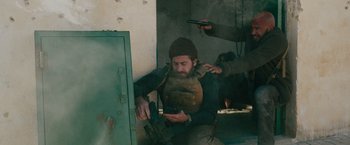 Movie still from “The Covenant” (2023), directed by Guy Ritchie – A man holding a cell phone while sitting on the ground; Medium shot, Over the shoulder angle