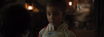 Movie still from “The Creator” (2023), directed by Gareth Edwards – A young boy wearing a white jacket with blue buttons; Close Up shot, Over the shoulder angle
