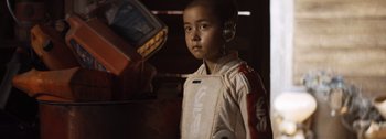 Movie still from “The Creator” (2023), directed by Gareth Edwards – A young boy in a costume is standing next to an electronic device; Close Up shot, Over the shoulder angle