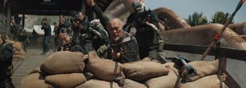 Movie still from “The Creator” (2023), directed by Gareth Edwards – An old man holding a gun while standing next to a pile of sacks; Medium shot, Over the shoulder angle