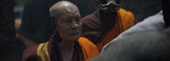 Movie still from “The Creator” (2023), directed by Gareth Edwards – An old man in a orange robe and a black mask; Close Up shot, Over the shoulder angle
