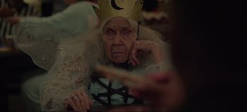 Movie still from “The Curse of Bridge Hollow” (2022), directed by Jeff Wadlow – An old woman wearing a paper crown and holding a cookie; Close Up shot, Over the shoulder angle