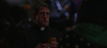 Movie still from “The Curse of Bridge Hollow” (2022), directed by Jeff Wadlow – A man wearing a priest's outfit holding a sandwich; Close Up shot, Over the shoulder angle