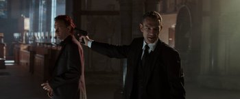 Movie still from “The Da Vinci Code” (2006), directed by Ron Howard – A man in a suit and tie pointing a gun at a woman; Medium shot, Over the shoulder angle