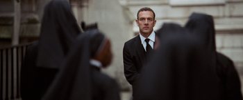 Movie still from “The Da Vinci Code” (2006), directed by Ron Howard – A man in a suit and tie standing in front of a group of people; Medium shot, Over the shoulder angle