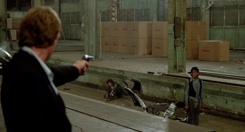 Movie still from “The Driver” (1978), directed by Walter Hill – A person is sitting on the ground with a gun pointed at him; Wide shot, Over the shoulder angle