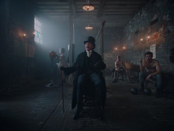 Movie still from “The Electrical Life of Louis Wain” (2021), directed by Will Sharpe – A man sitting in a chair in a room with other people; Wide shot, Low angle