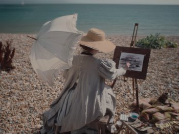 Movie still from “The Electrical Life of Louis Wain” (2021), directed by Will Sharpe – A woman with an umbrella painting a picture on the beach; Medium shot, High angle