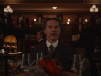 Movie still from “The Electrical Life of Louis Wain” (2021), directed by Will Sharpe – A man sitting at a dinner table with wine glasses in front of him; Medium shot, Over the shoulder angle