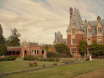 Movie still from “The Electrical Life of Louis Wain” (2021), directed by Will Sharpe – People are sitting in the grass outside of a castle like building; Extreme Wide shot, Low angle