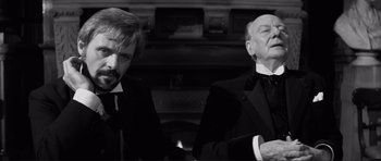 Movie still from “The Elephant Man” (1980), directed by David Lynch – A man and a man in a suit standing next to each other in front of a fireplace; Medium shot, Over the shoulder angle