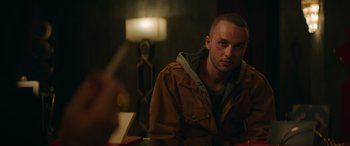 Movie still from “The Enforcer” (2022), directed by Richard Hughes – A man sitting at a table in front of a lamp; Close Up shot, Over the shoulder angle