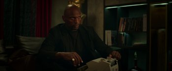 Movie still from “The Enforcer” (2022), directed by Richard Hughes – A man sitting in a chair looking at the camera; Medium shot, Low angle