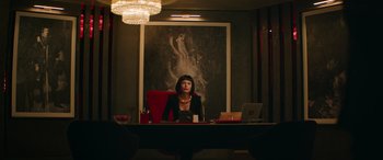 Movie still from “The Enforcer” (2022), directed by Richard Hughes – A woman sitting at a desk in front of a painting; Medium shot, Low angle