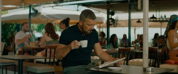 Movie still from “The Enforcer” (2022), directed by Richard Hughes – A man sitting at a table with a cup of coffee and looking at a tablet; Medium shot, High angle