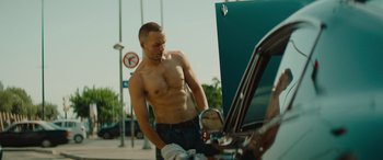 Movie still from “The Enforcer” (2022), directed by Richard Hughes – A shirtless man standing next to an open car hood; Medium shot, Low angle