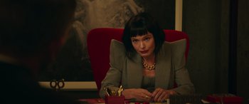 Movie still from “The Enforcer” (2022), directed by Richard Hughes – A woman sitting at a table in a red chair; Medium shot, Over the shoulder angle