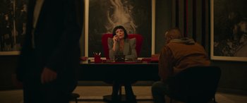 Movie still from “The Enforcer” (2022), directed by Richard Hughes – A woman sitting at a table talking on a phone; Medium shot, Over the shoulder angle