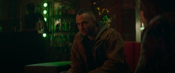 Movie still from “The Enforcer” (2022), directed by Richard Hughes – A man sitting in front of a bunch of bottles in a room; Medium shot, Low angle