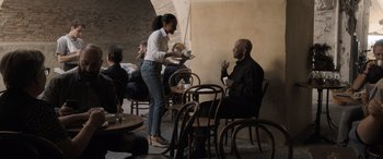 Movie still from “The Equalizer 3” (2023), directed by Antoine Fuqua – A woman standing at a table with a plate in front of a man; Wide shot, Over the shoulder angle