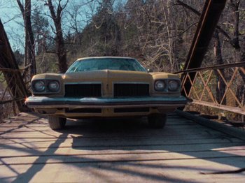 Movie still from “The Evil Dead” (1981), directed by Sam Raimi – An old car is parked on the side of a bridge; Wide shot, Low angle