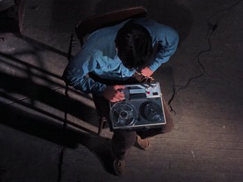 Movie still from “The Evil Dead” (1981), directed by Sam Raimi – A man sitting in a chair holding a tape recorder; Wide shot, Overhead angle