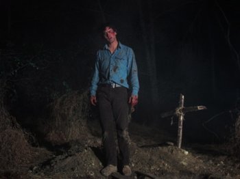 Movie still from “The Evil Dead” (1981), directed by Sam Raimi – A man standing next to a cross in the dark; Wide shot, Low angle