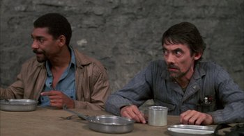 Movie still from “Day of the Dead” (1985), directed by George A. Romero – Two men sitting at a wooden table with plates and cups; Medium shot, High angle