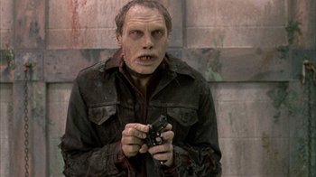 Movie still from “Day of the Dead” (1985), directed by George A. Romero – A man holding a cell phone while wearing a zombie costume; Medium shot, Low angle
